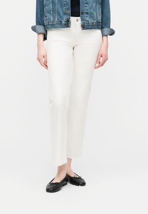 Woman wearing white straight-leg pants, black ballet flats, and a rolled-up denim jacket against a plain white background.