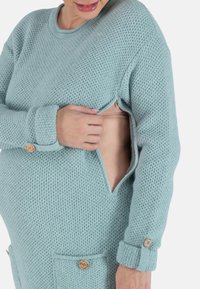 Pull en tricot bleu clair avec un motif texturé, comprenant une fermeture éclair à l'avant et des accents en bois sur les poignets et les poches.
