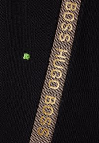 Schwarzer Stoff mit einem braunen gewebten Band, auf dem "BOSS HUGO BOSS" in Gold steht, sowie einem kleinen grünen Stickdetail.