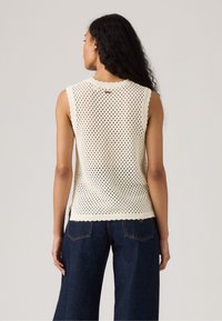 Maglione senza maniche a maglia color crema con un motivo aperto a nido d'ape, bordi ondulati e scollatura rotonda. Abbinato a pantaloni larghi in denim scuro.