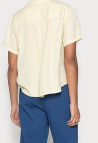 Chemise jaune à manches courtes avec une coupe décontractée, dotée d'un panneau arrière et d'un léger éclat, associée à un pantalon bleu. Tissu lisse et léger.