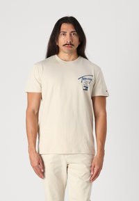 Beige kortærmet t-shirt lavet af bomuld, med et blåt logo på den øverste venstre bryst med en tegning. Enkelt design, rund hals.