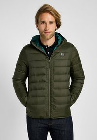 Olivgrön puffjacka med dragkedja framtill, horisontellt quiltad design och en teal-fodrad huva. Har en liten logotyp på bröstet.