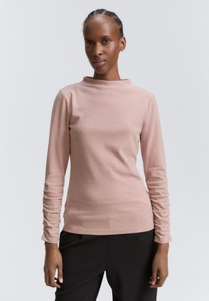 Donna con capelli intrecciati che indossa un top rosa chiaro a maniche lunghe e pantaloni neri, in piedi contro uno sfondo grigio semplice.