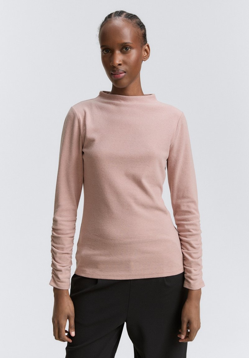 Kobieta z zaplecionymi włosami, ubrana w różową bluzkę z długim rękawem i czarne spodnie, stojąca na jednolitym szarym tle.