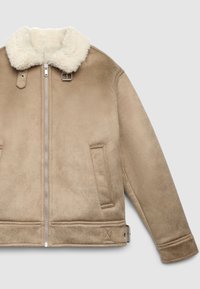 Giacca beige in shearling con colletto in pelliccia bianca, chiusura frontale con zip, due tasche laterali e dettaglio con fibbia sul colletto. Texture morbida.