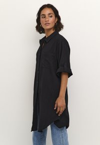 Chemise noire à col, en tissu texturé, avec une poche poitrine unique et des manches retroussées, associée à un jean bleu.