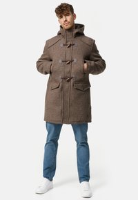 Brauner Woll-Dufflecoat mit Kapuze, Knebelverschlüssen, zwei Fronttaschen und strukturierter Oberfläche. Kombiniert mit hellblauen Jeans und weißen Sneakers.