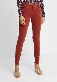 Korsett-jeans i rödbrun kordflossa, med framfickor och en figurnära silhuett. Matchas med beige klacksandaler med remmar.