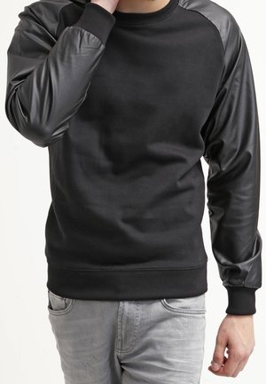 Mann trägt schwarzen Sweatshirt mit Ledermanschetten und hellgraue Jeans, Hand berührt das Gesicht, beschnitten auf Kinn und Hüften.