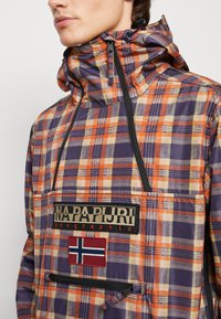 Person iført en orange, lilla og cremefarvet plaid hættetrøje med frontlommer og Napapijri logo patch med norsk flag.