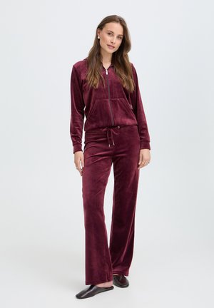 Femme debout portant une veste zippée en velours bordeaux et un pantalon à cordon assorti avec des chaussons noirs sur un fond uni.