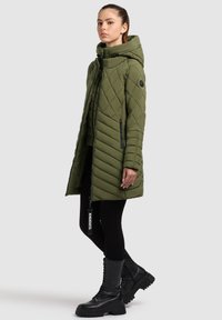 Cappotto lungo trapuntato verde oliva con cappuccio, dotato di tasche con zip e un'etichetta con logo. Abbinato a leggings neri e stivali da combattimento con lacci.