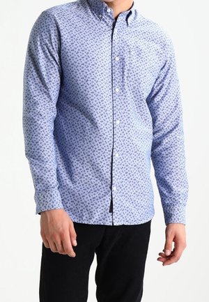 Mann trägt ein hellblaues langärmeliges Hemd mit kleinem dunklen Muster und schwarze Hose, stehend vor einem schlichten, hellen Hintergrund.