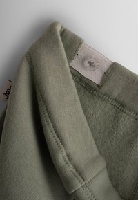 Svetlo zelena kapuca s mekim tekstilom. Vidljiva šavna detalja i plastični gumb blizu oznake. Oznaka glasi "Jo!"