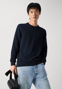 Marineblauer Pullover mit strukturiertem Strickdesign, Rundhalsausschnitt und gerippten Bündchen, kombiniert mit hellblauen Jeans und einer schwarzen Tasche.