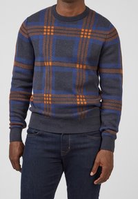 Pull gris avec un motif à carreaux bleu et orange, poignets et ourlet côtelés, fabriqué en tissu tricoté doux. Associé à un jean foncé.