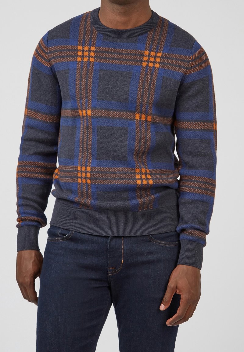 Pull gris avec un motif à carreaux bleu et orange, poignets et ourlet côtelés, fabriqué en tissu tricoté doux. Associé à un jean foncé.