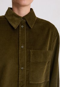 Chemise en velours côtelé vert foncé avec une coupe légèrement surdimensionnée, dotée d'un col à boutonnière, d'une poche frontale et d'une texture à côtes verticales.