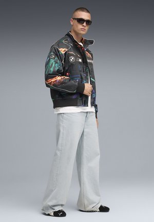 Schwarze Bomberjacke mit bunten abstrakten Grafiken, weißen Akzenten und BMW-Logo. Kombiniert mit hellblauen lässigen Jeans und schwarzem Schuhwerk.