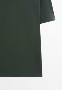 Chemise à manches courtes vert foncé montrant le côté et l'ourlet inférieur sur fond blanc.