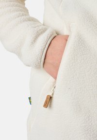 Weiße Fleecejacke mit strukturierter Oberfläche, ausgestattet mit einer seitlichen Reißverschlusstasche und einem kleinen Lederetikett. Die Ärmelbündchen sind gerippt für eine optimale Passform.