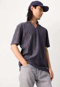 Polo shirt grigio a maniche corte realizzato in tessuto testurizzato con un delicato motivo a onde, caratterizzato da un colletto a V e una vestibilità rilassata. Abbinato a jeans grigi chiari.