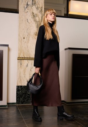 Pull noir court, jupe midi marron, bottines en cuir noir, et un sac à main noir avec un accent doré. Textures lisses ; design minimal.