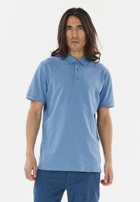 Vaaleansininen polo-paita, joka on valmistettu pehmeästä puuvillasta. Siinä on kolme nappia ja lyhyet hihat. Yksinkertainen muotoilu, jossa on hillitty logo rinnassa.