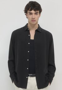 Junger Mann mit dunklem Haar trägt ein aufgeknöpftes schwarzes Hemd über einem schwarzen Tanktop, beigen Hosen und einem schwarzen Gürtel und steht vor einem einfarbigen Hintergrund.