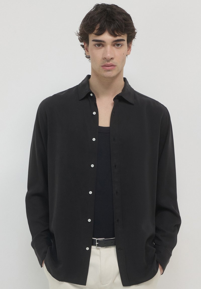 Junger Mann mit dunklem Haar trägt ein aufgeknöpftes schwarzes Hemd über einem schwarzen Tanktop, beigen Hosen und einem schwarzen Gürtel und steht vor einem einfarbigen Hintergrund.