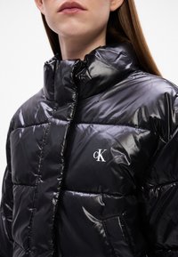 Giacca a vento nera lucida con colletto alto, dotata di un piccolo logo bianco "ck", design trapuntato e tasche laterali.