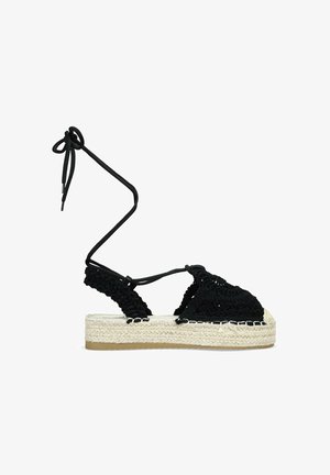 Espadrilles de crochet negro con lazos en el tobillo, con una suela intermedia de yute tejido y suela de goma, que destacan por su parte superior texturizada y diseño plano de plataforma.
