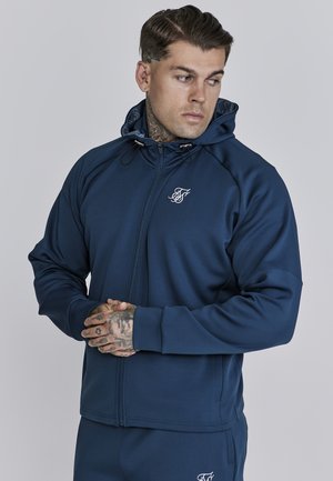 Sudadera con cremallera color navy, con capucha ajustable, tela texturizada y un pequeño logo blanco en el pecho. Presenta mangas raglán y puños acanalados.