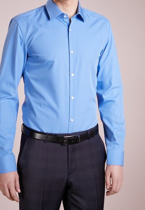 Mann trägt ein tailliertes hellblaues Hemd mit Knöpfen, schwarzen Gürtel und dunkle gemusterte Stoffhose vor einfachem Hintergrund.