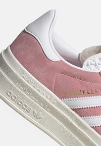 Zapatillas Adidas Gazelle de ante rosa con rayas blancas, tapón en el talón, suela texturizada y el logo dorado "GAZELLE" en el lateral.