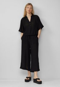 Schwarzes zweiteiliges Outfit mit einem kurzärmeligen, kragenlosen Top mit Knöpfen und lockeren, cropped Hosen. Aus strukturiertem Stoff, minimales Design.