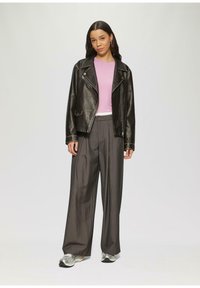 Zwarte leren motorjack, roze top met korte mouwen, brede gestreepte broek, zilveren sneakers, staand tegen een lichtgrijze achtergrond.