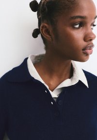 Marineblauer Wollpullover mit Kragen über einem weiß-grau gestreiften Hemd, das eine geknöpfte Vorderseite und eine strukturierte Stoffoberfläche aufweist.