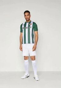 Grønn og hvit stripete fotballdrakt med korte ermer, med logo og teksten "TRUST IT." Kombinert med hvite shorts og sokker, samt svarte detaljer på fottøyet.