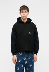 Μαύρο γιλέκο με φερμουάρ και κουκούλα, μπροστινές τσέπες και μικρό λογότυπο. Συνδυασμένο με μπλε τζιν που έχουν σχέδιο yin-yang.