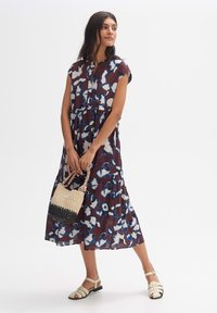 Blumenmusterkleid mit einem gestuften Design in Burgunderrot, Blau und Creme; kurze Ärmel; kombiniert mit einer gewebten schwarzen und cremefarbenen Handtasche sowie flachen Sandalen.