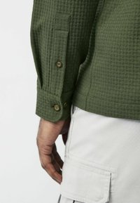 Groene shirt met textuur, lange mouwen en knoopmanchetten met bruine knopen, gecombineerd met lichte cargobroek.