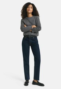 Grå kabelstickad tröja, mörkblå raka jeans, svarta skor, stående pose. Avslappnad outfit med texturerad stickning och denim.