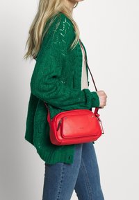 Czerwona skórzana torba crossbody z dwoma przednimi kieszeniami na zamek i odpinanym paskiem, noszona na zielonym swetrze dzianinowym i niebieskich dżinsach.