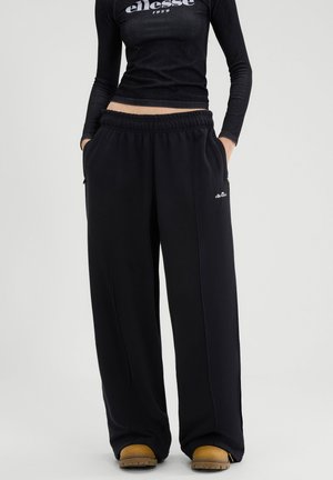 Person iført sort ellesse langærmet croppet top og løse sorte ellesse bukser, stående med hænderne i lommerne og brune støvler synlige.