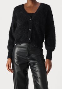 Czarny, puszysty kardigan z dekoltem w serek i długimi rękawami. Posiada trzy dekoracyjne guziki z przodu oraz krótki fason. Połączony z czarnymi spodniami z sztucznej skóry.