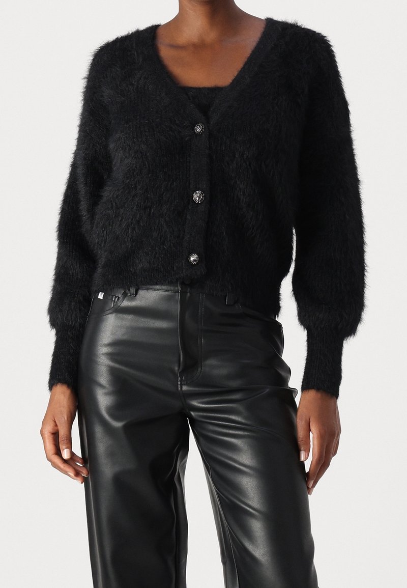 Czarny, puszysty kardigan z dekoltem w serek i długimi rękawami. Posiada trzy dekoracyjne guziki z przodu oraz krótki fason. Połączony z czarnymi spodniami z sztucznej skóry.