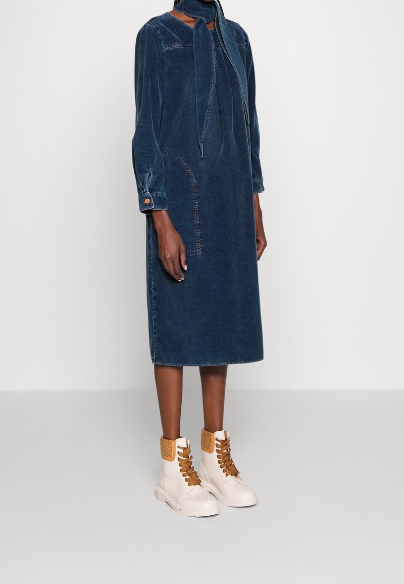 Robe en denim, bleu marine, midi avec un col à nouer, des manches longues et des détails brodés. Assortie à des bottes blanches avec des accents marron.