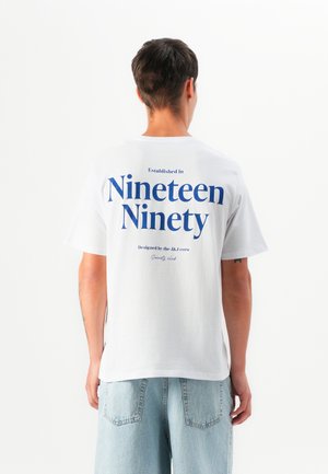 T-shirt en coton blanc avec un grand texte bleu au dos : "Établi en mille neuf cent quatre-vingt," avec des détails plus petits en dessous. Coupe décontractée.